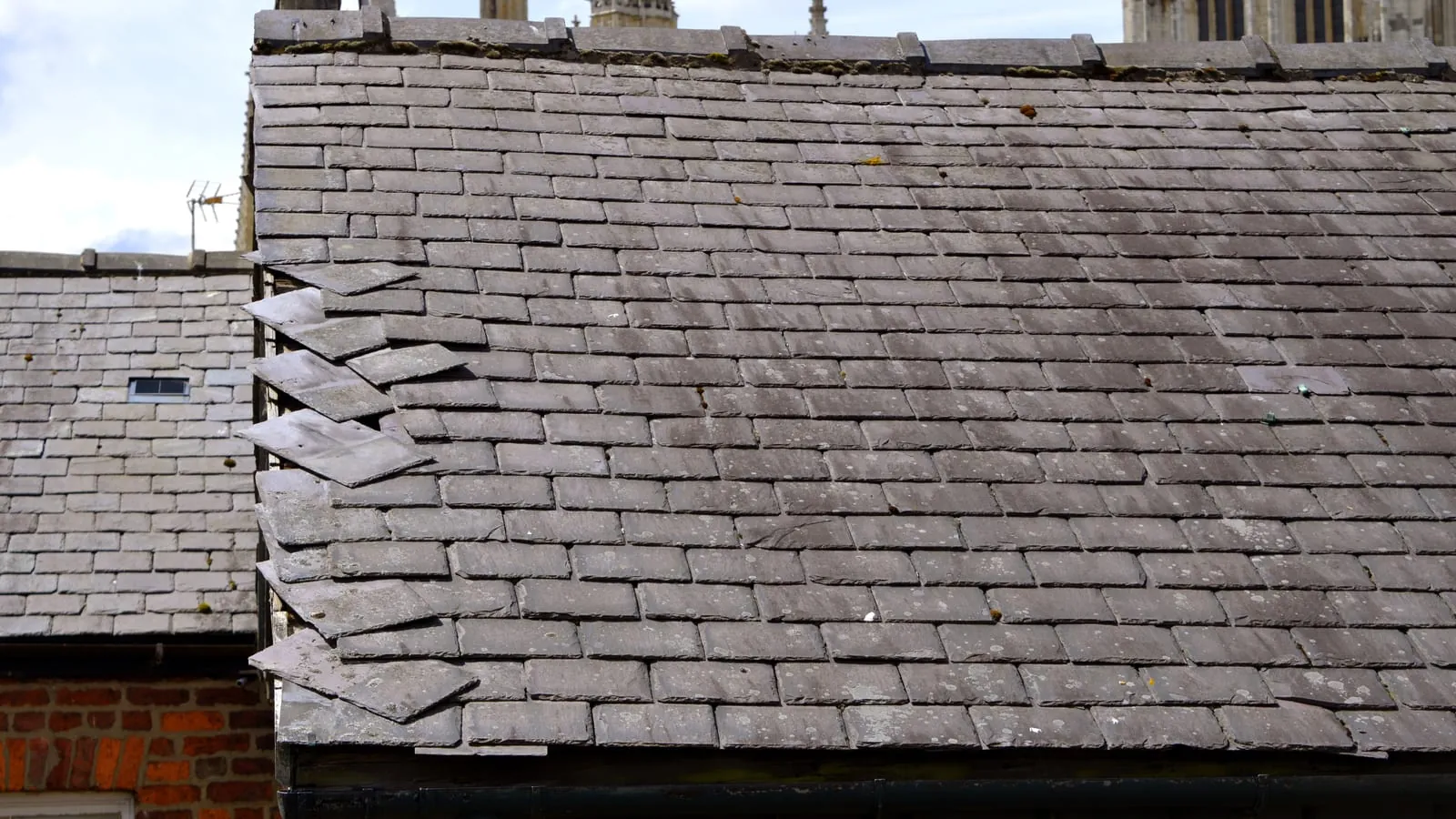 A run of clay tiles slipped out of course on a weathered roof above a brick gable